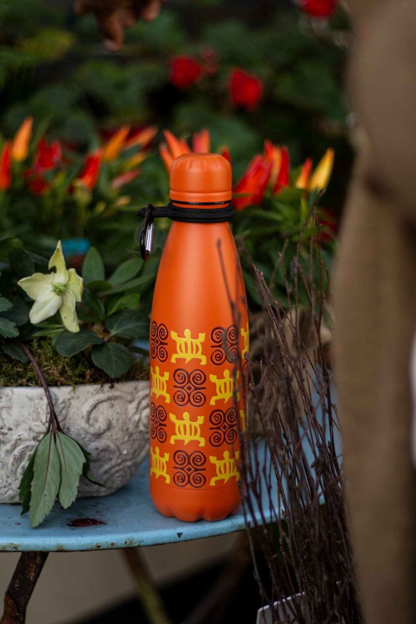 orange water bottle with adrinka symbols on a table with colourful flowers in the background.