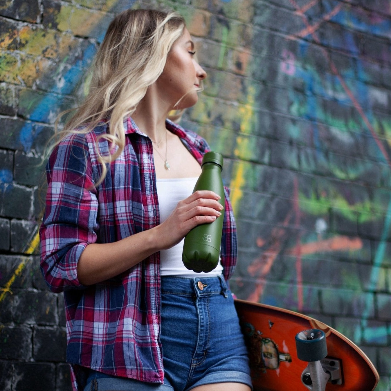Lady with skateboard and stands at a wall with a green water bottle in hand. looks into the distance.