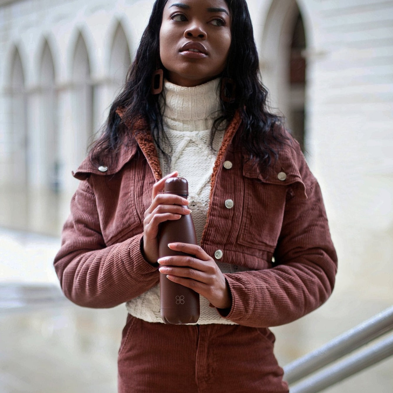 Lady in brown corduroy jacket holds a matching brown stainless steel bottle.