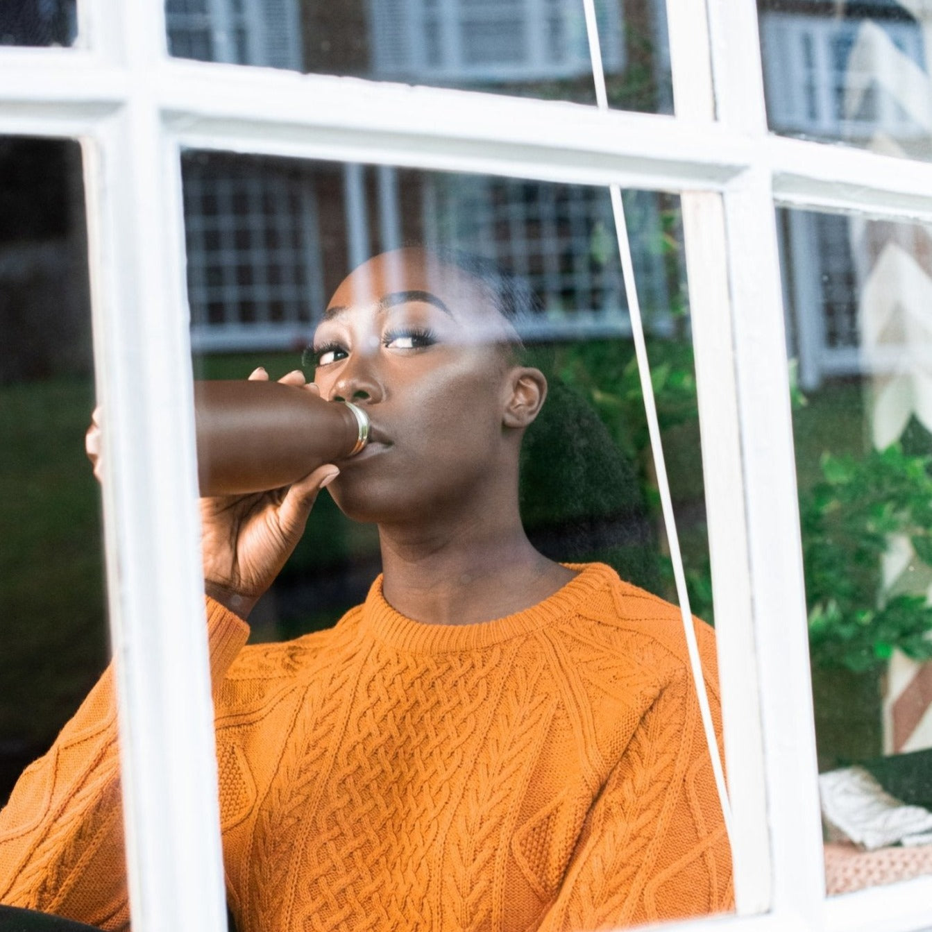 Lady in a sun orange jumper sits in front of a windoe drinking from a brown steel water bottle